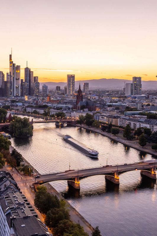 Blick auf den Main, die Frankfurter Skyline und den Kaiserdom vom Dach des Lindner Hotel Frankfurt Main Plaza bei Abenddämmerung.