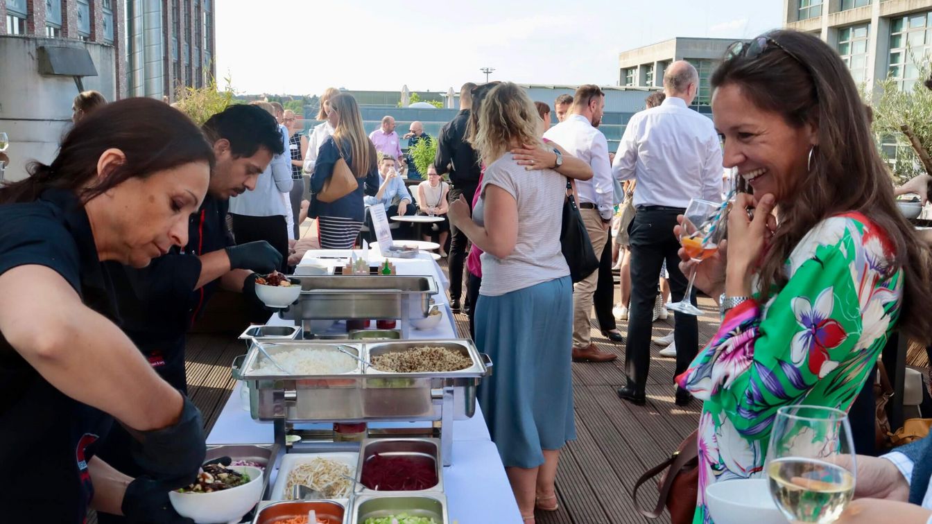 Veranstaltungsteilnehmende bei blauem Himmel und Sonnenschein am Buffet des Get-Togethers des FRM MICE Networks auf der Dachterrasse des Belle Roofgarden.